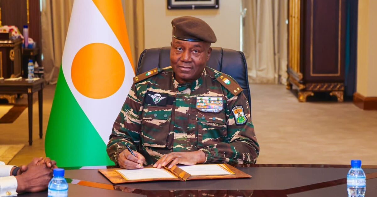 Niger military ruler Abdourahmane Tchiani seated beside a Niger flag.