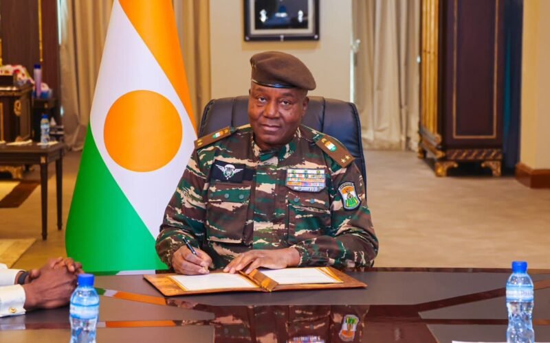 Niger military ruler Abdourahmane Tchiani seated beside a Niger flag.