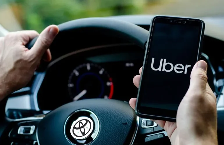 A driver holding a phone displaying the Uber app logo while seated behind a steering wheel