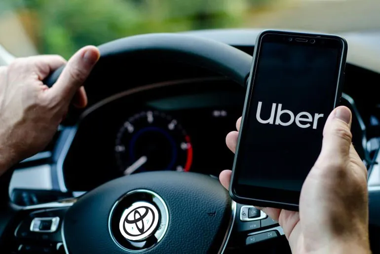 A driver holding a phone displaying the Uber app logo while seated behind a steering wheel