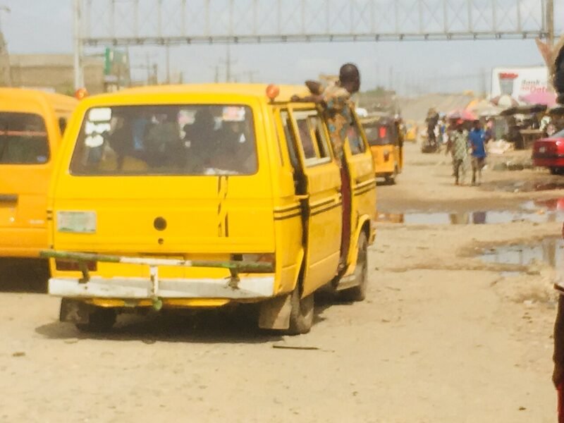 A yellow danfo minibus seen from behind on a Lagos road, with a passenger boarding.