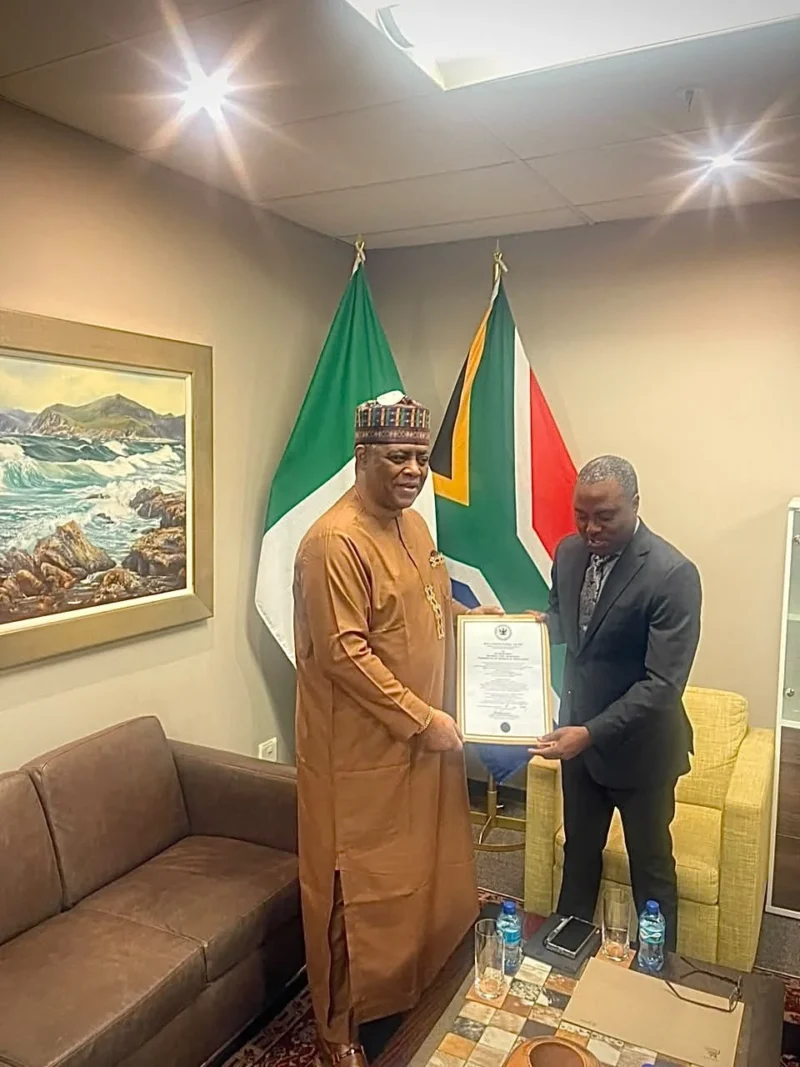 Femi Fani-Kayode, in agbada and cap, presents his credentials to a South African protocol official between the Nigerian and South African flags.