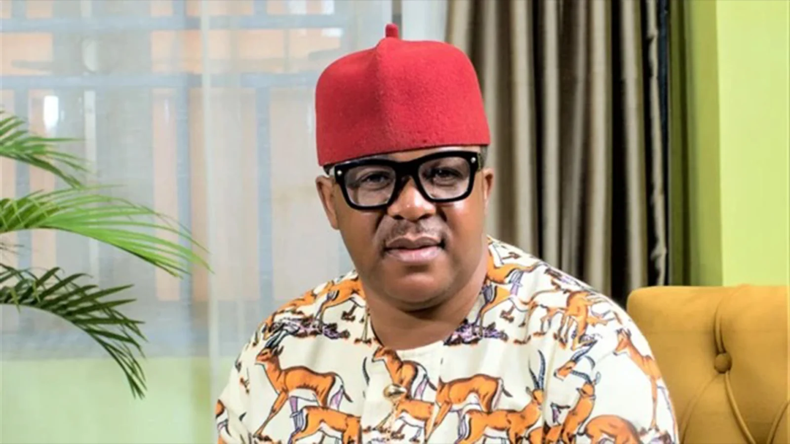 Nollywood actor Francis Duru wearing a red cap and patterned dress, seated indoors