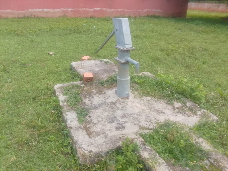 A hand-operated borehole water pump mounted on a concrete platform beside a building in Oyo State, Nigeria.