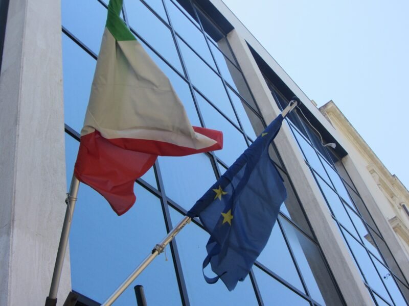 The Italian and European Union flags flying outside a glass government building