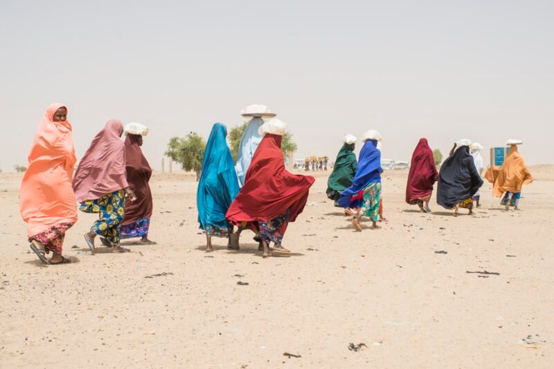 Women carrying bundles on their heads walking across a dusty landscape in West Africa, in a scene from displaced displacement-affected communities.