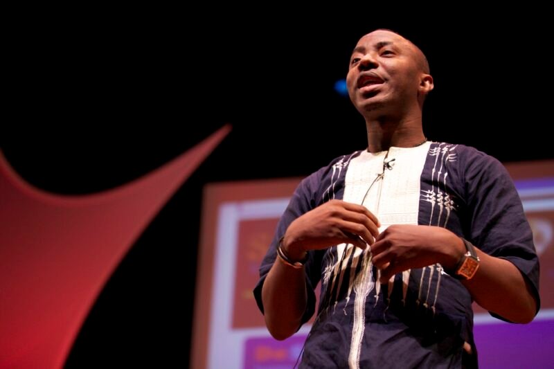 Omoyele Sowore, in a dark embroidered shirt, speaks on a stage in front of a projection screen.