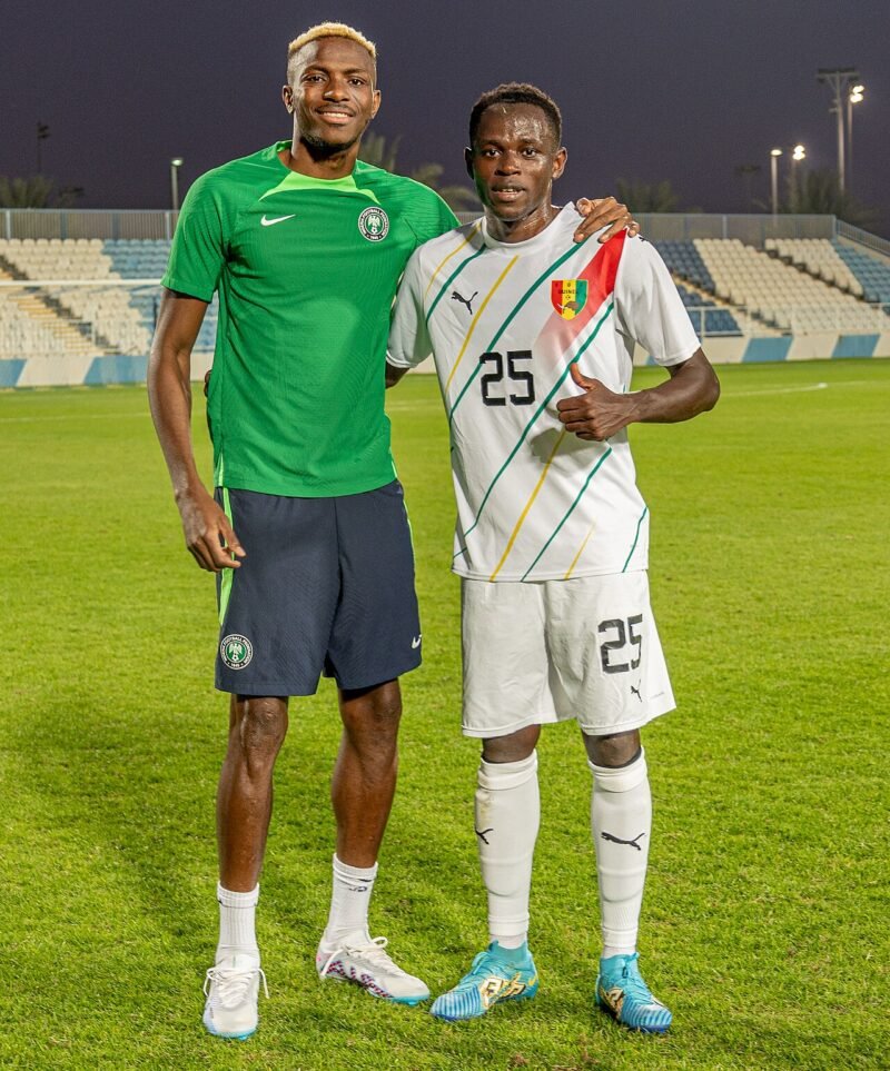 Victor Osimhen in a green Nigeria shirt stands beside Guinea player Facinet Conte on a football pitch.