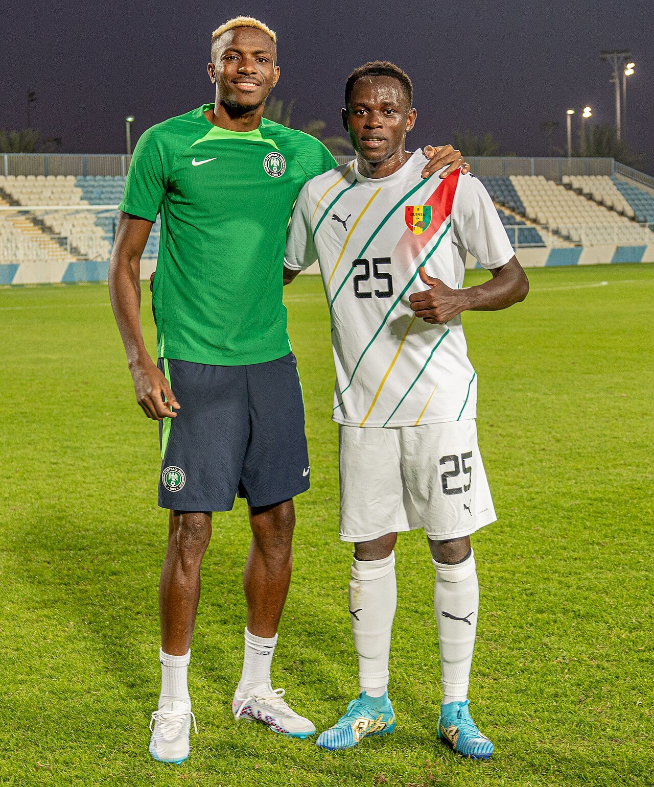 Victor Osimhen in a green Nigeria shirt stands beside Guinea player Facinet Conte on a football pitch.