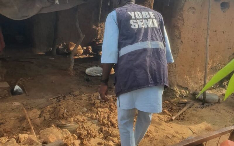 A worker wearing a Yobe SEMA vest stands beside an earthen shelter and debris.