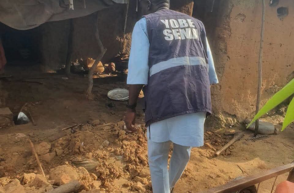A worker wearing a Yobe SEMA vest stands beside an earthen shelter and debris.